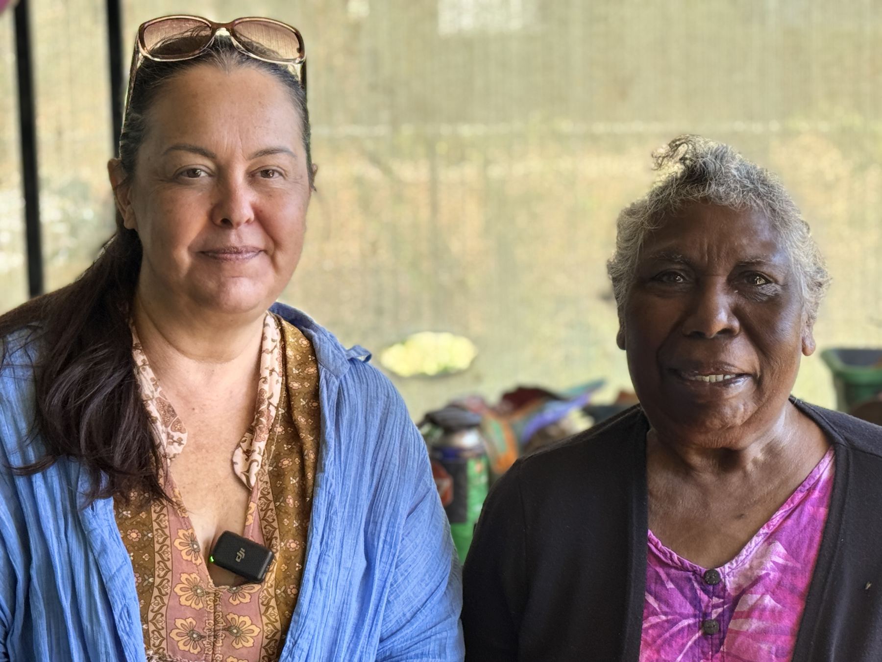 Two women smiling together at a community gathering, one wearing a lapel microphone