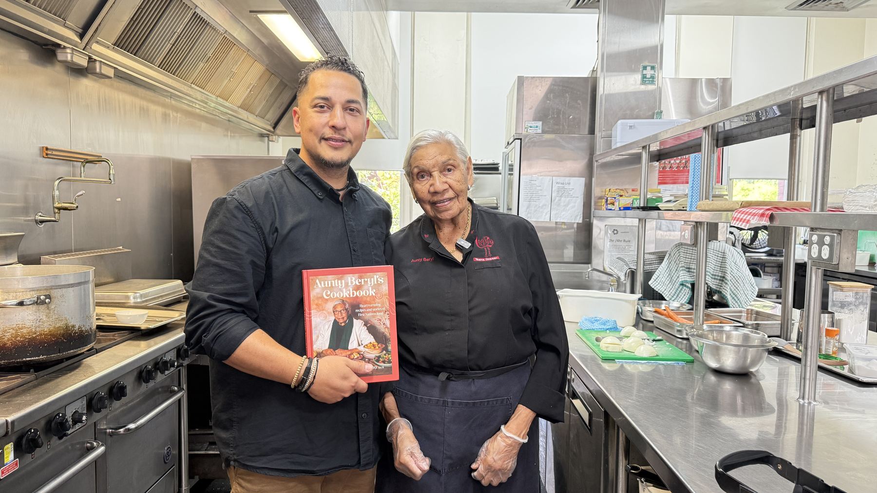 Levi-Joel Tamou and Aunty Beryl Van-Oploo in the NCIE kitchen holding Aunty Beryl's Cookbook